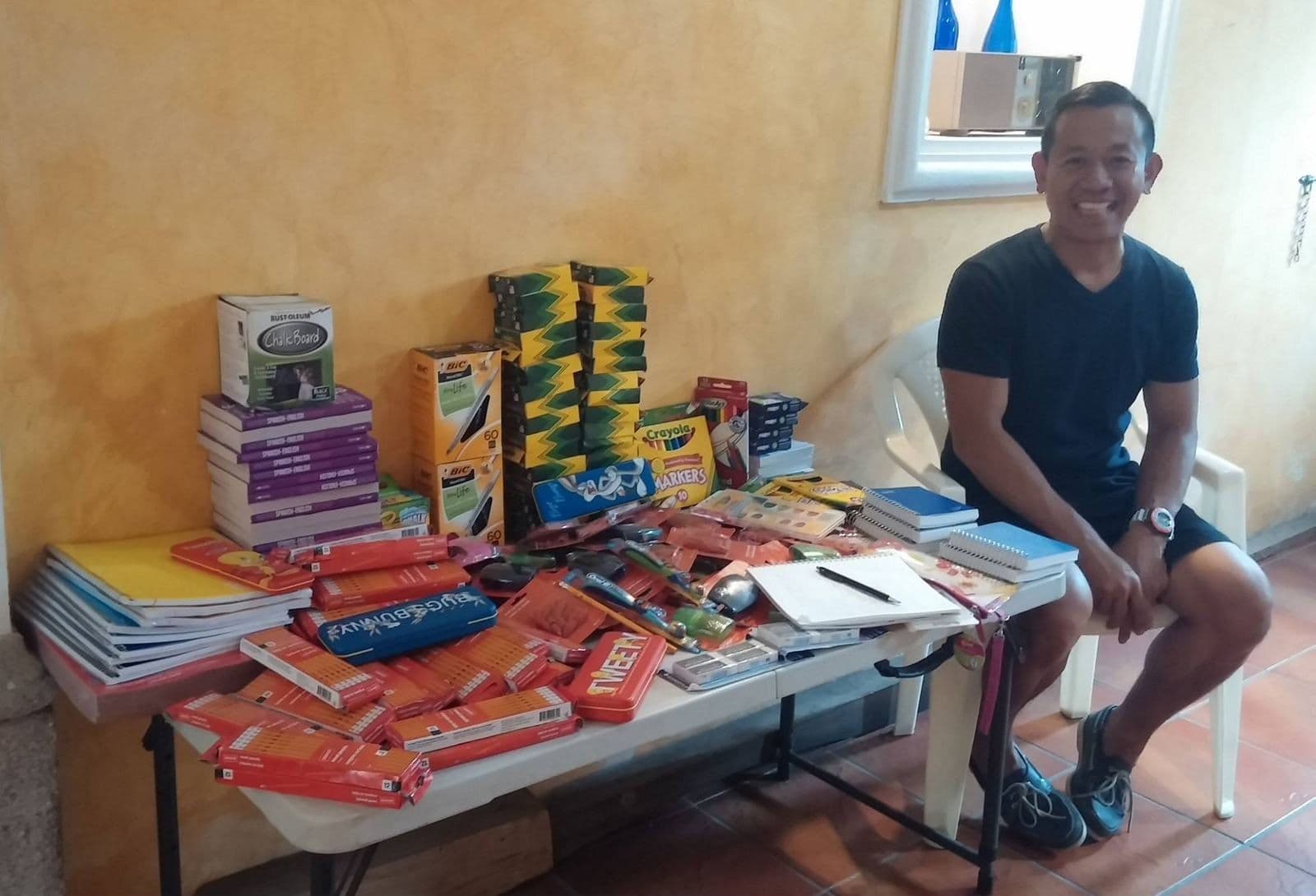 A donor sits beside a table of notebooks, crayons and markers he brought for the children