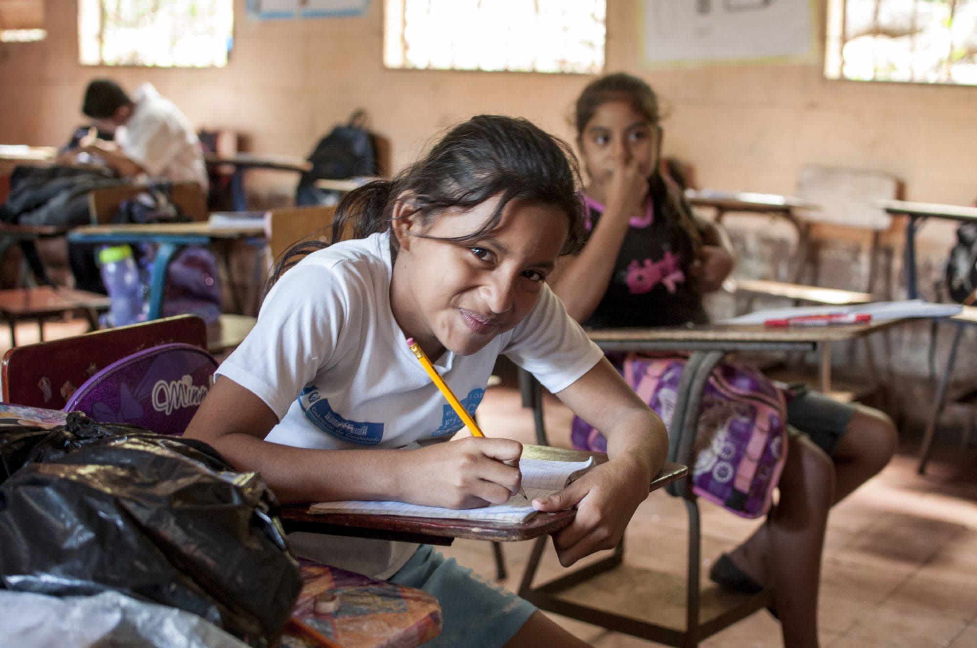 A girl writes in her notebook and glances playfully at the camera in a Las Delicias classroom