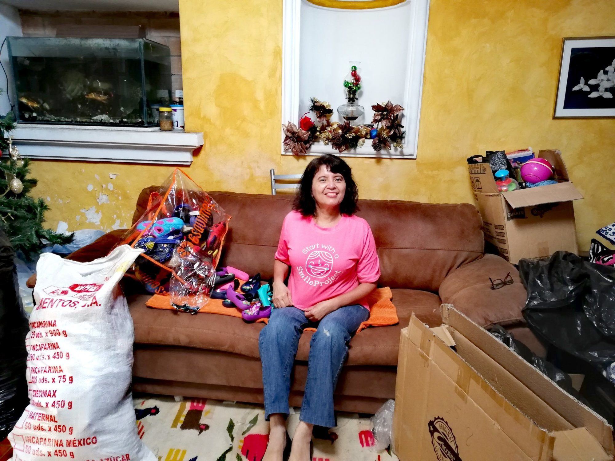 Amalia, in a project T-shirt, sits in her living room among donated shoes, boxes and bags