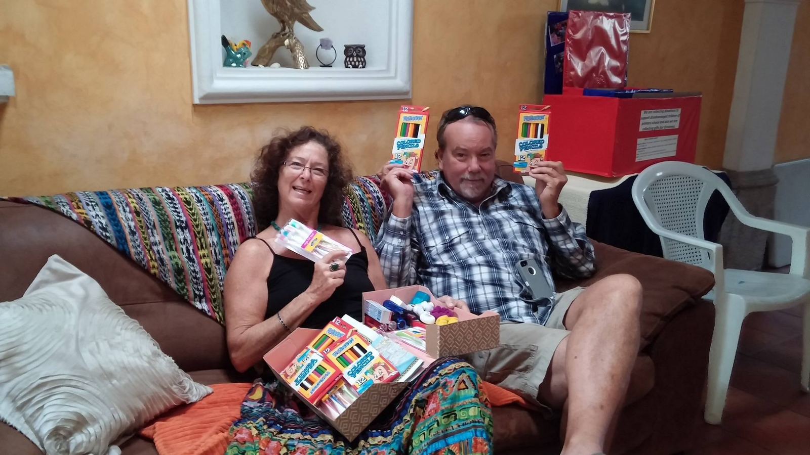 Two donors hold up colored pencils and toothbrushes on a sofa draped with a Guatemalan textile
