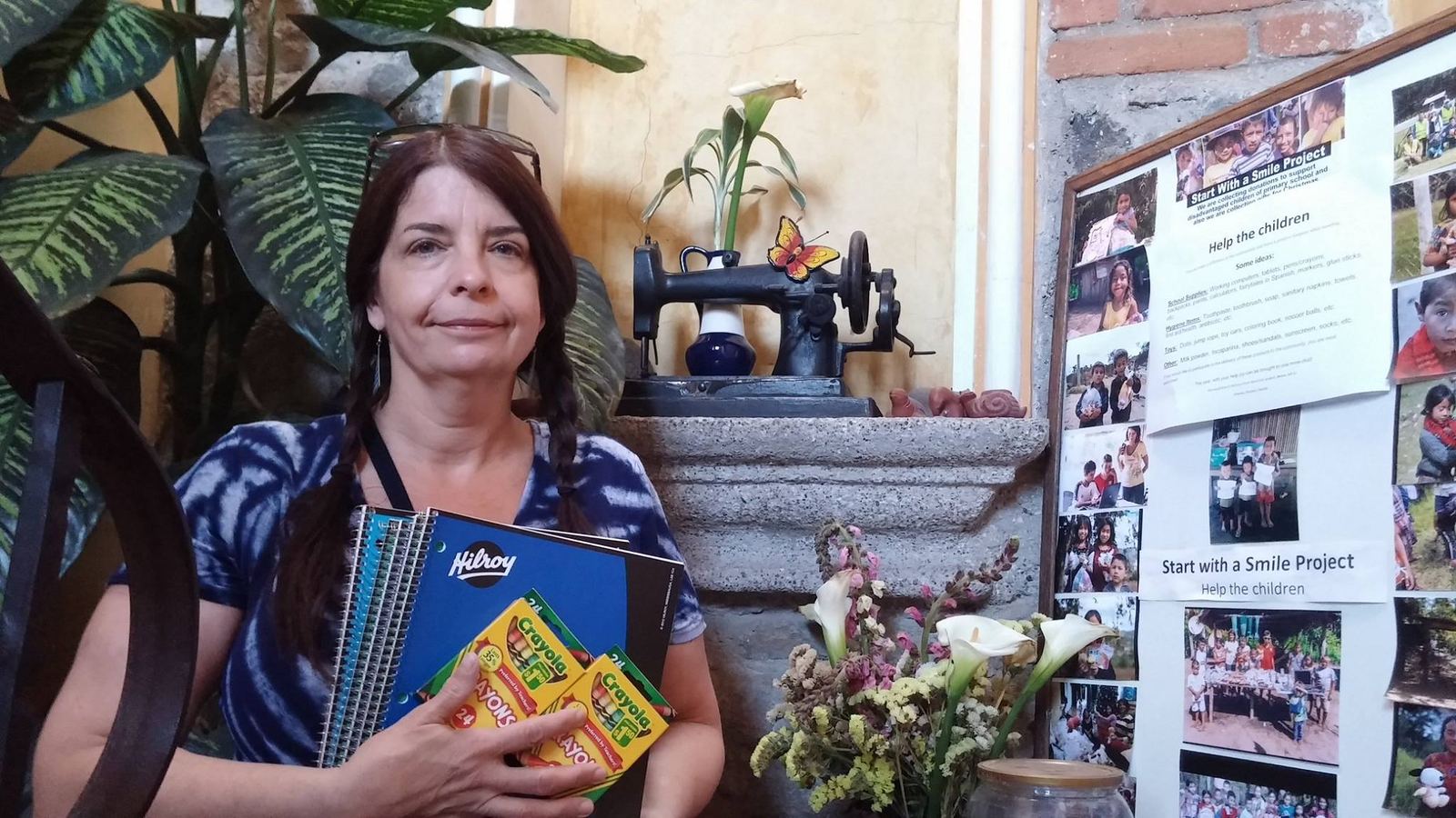 A donor holds notebooks and crayons beside the project's photo board and an antique sewing machine