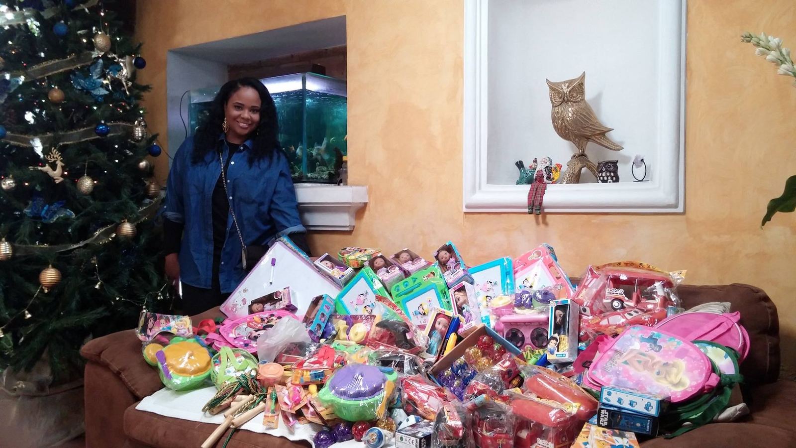 A donor stands by a sofa piled with dolls, toy cars, backpacks and other toys next to a Christmas tree
