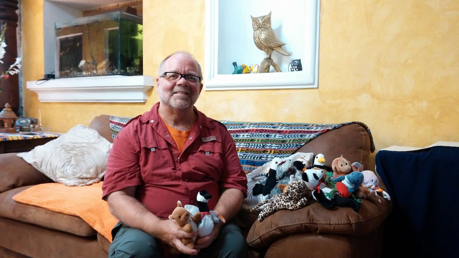 A smiling donor on a sofa draped with a woven textile holds two stuffed animals, with more beside him