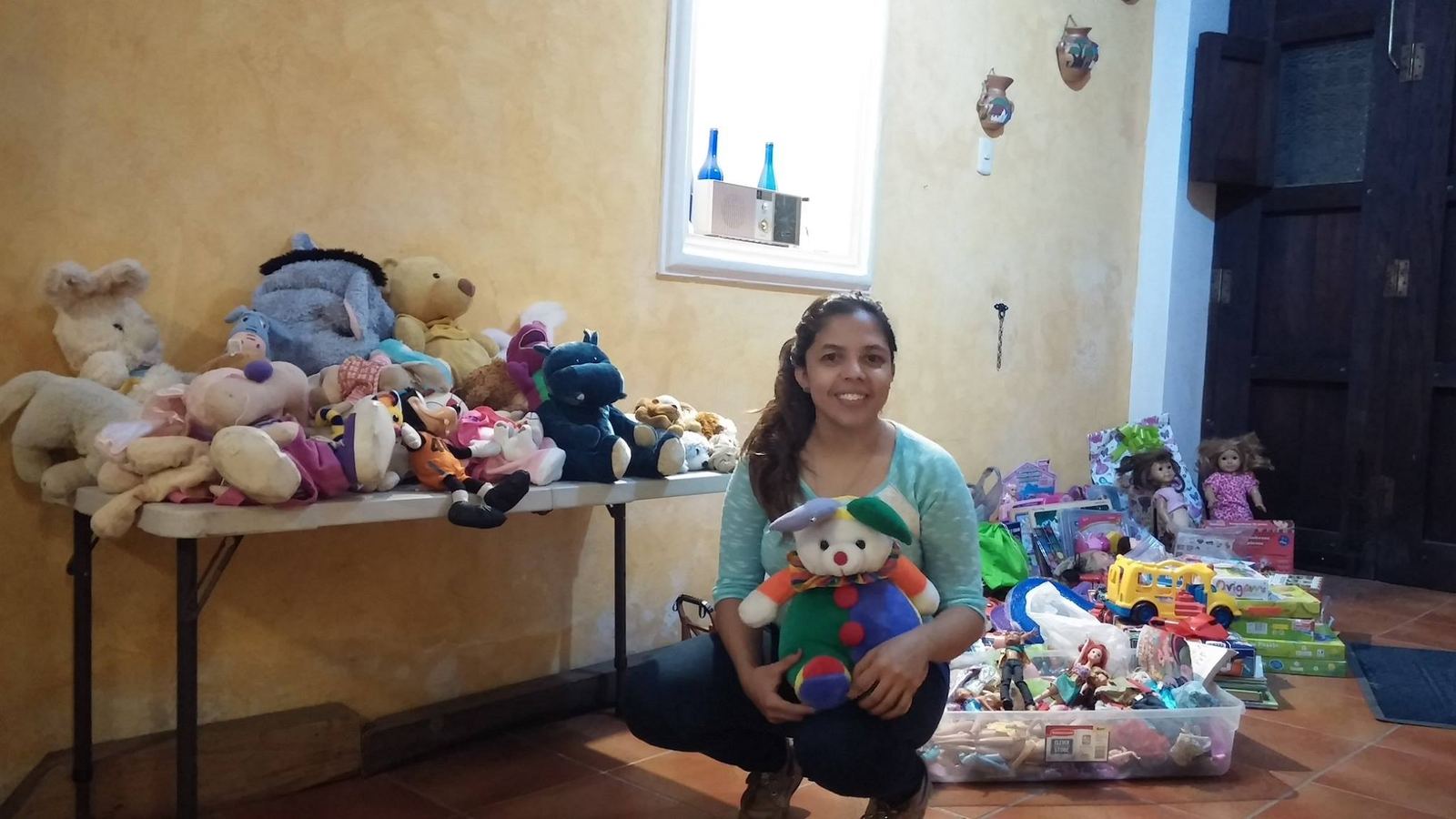 A donor hugs a teddy bear beside a table full of stuffed animals and a bin of dolls