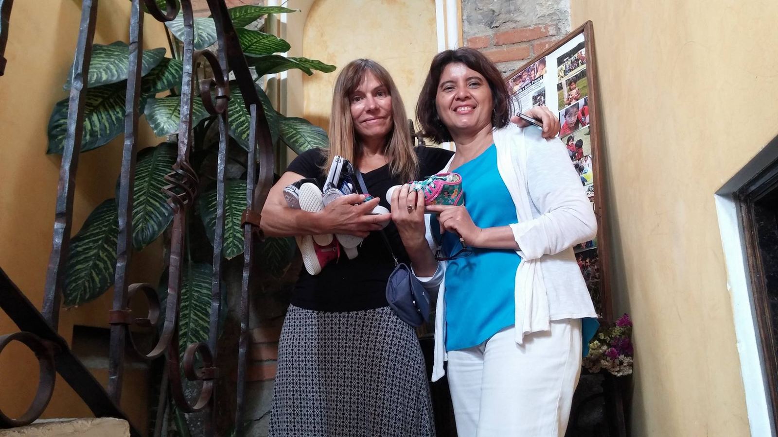 Two women smile by a wrought-iron railing holding children's sneakers donated to the project