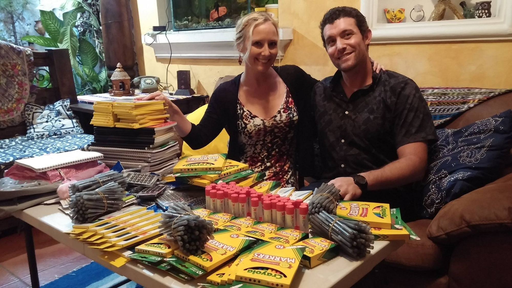 Two donors smile behind a table piled with crayons and school supplies in a home in Antigua