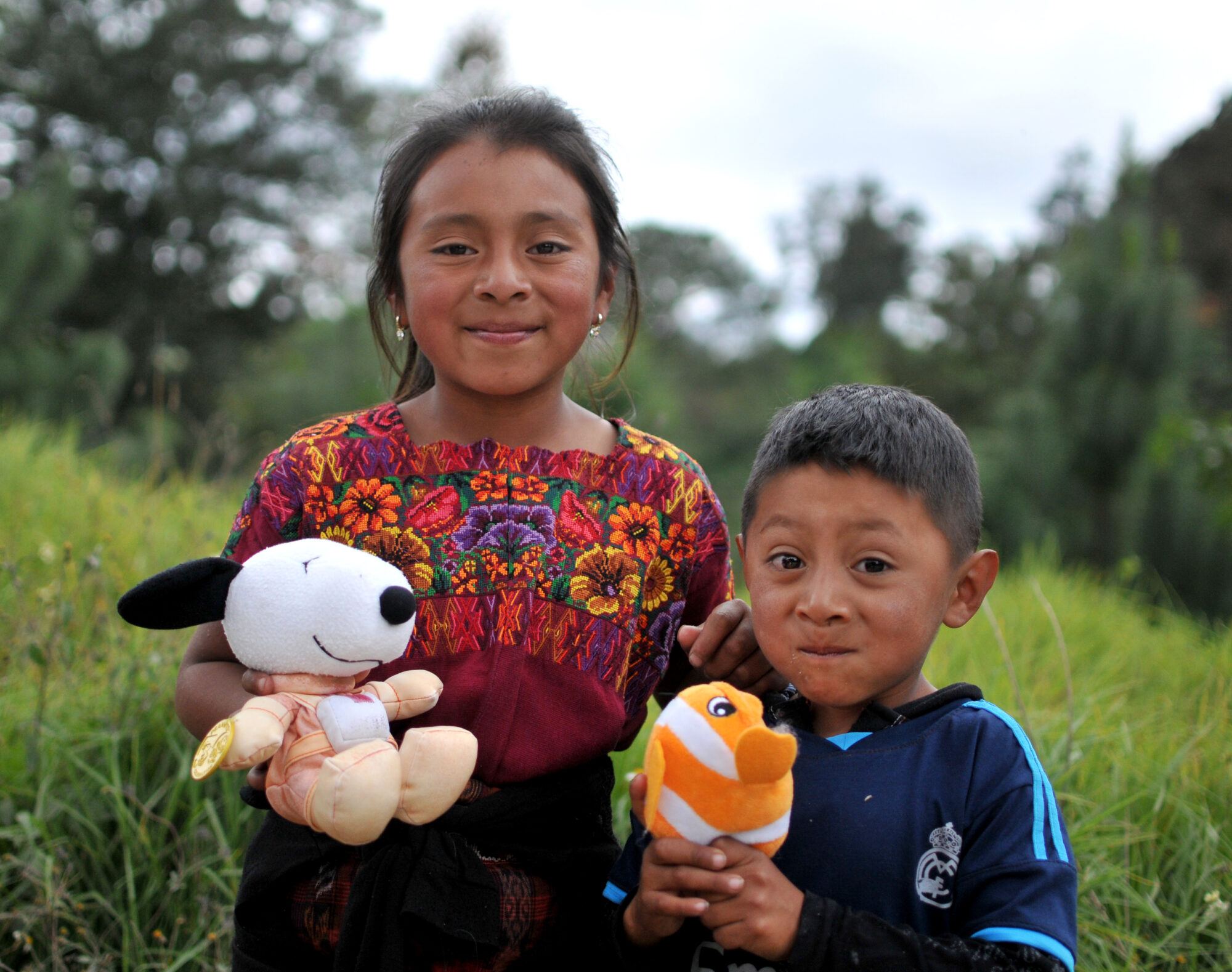 A girl in a traditional huipil and a small boy pose with their Snoopy and clownfish plush toys