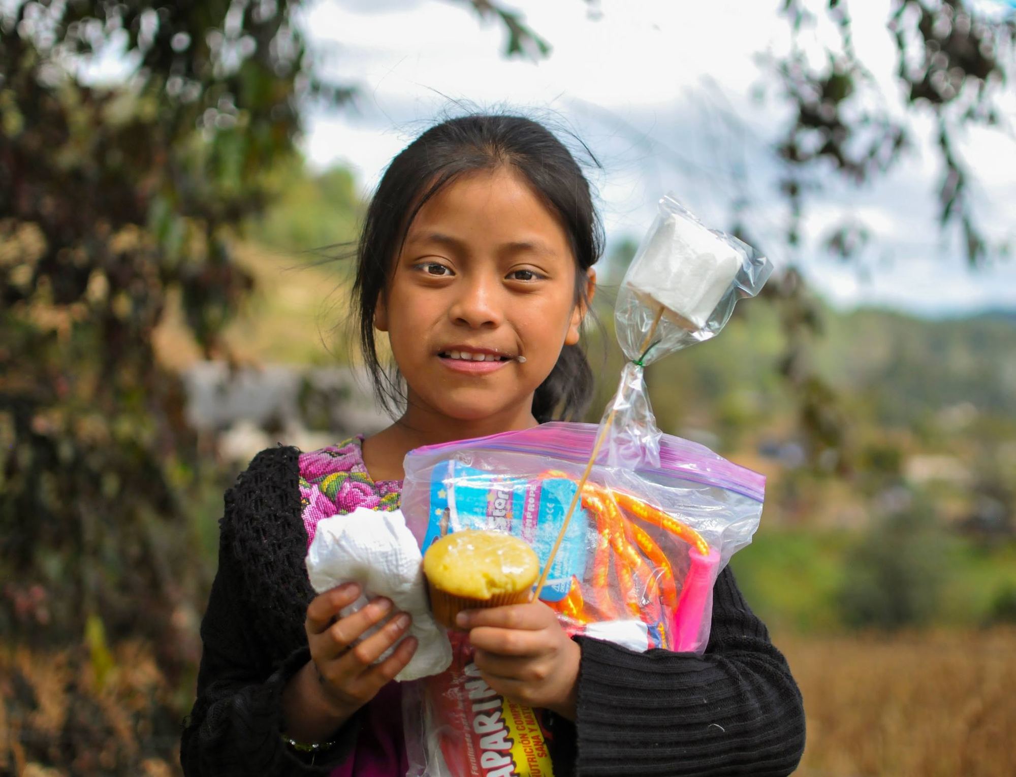 Una niña con huipil sostiene su bolsa de regalo, un pastelito y un malvavisco, con el valle del altiplano detrás