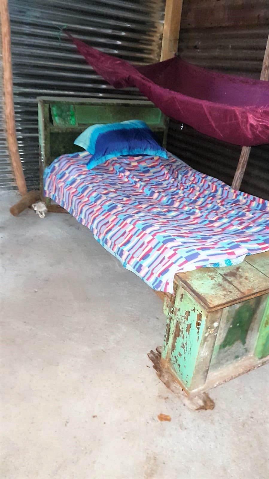 A made bed with a colorful sheet and a purple hammock inside a metal-sheet home with a cement floor