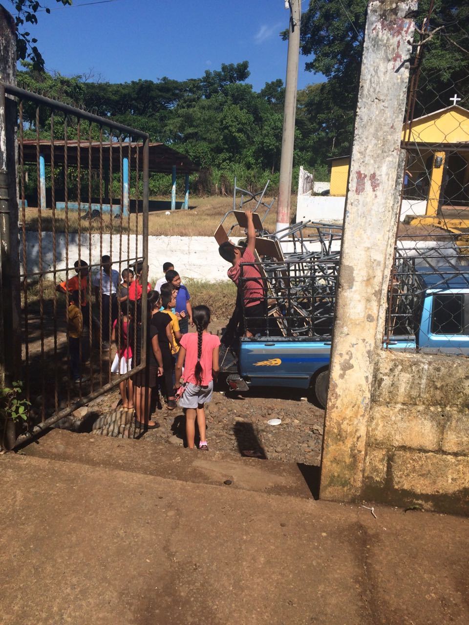 A young man unloads desks from a light blue pickup as a group of children waits at the school gate
