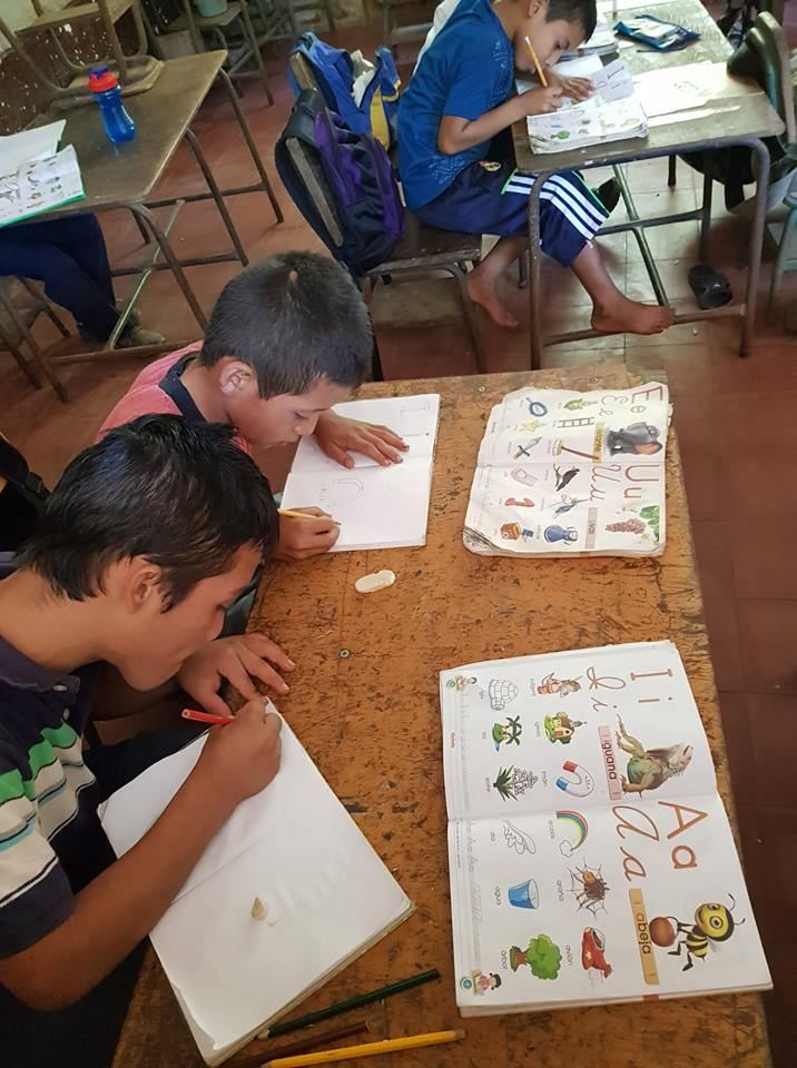 Two boys write in their notebooks next to reading primers open to the letters A, I, E and U