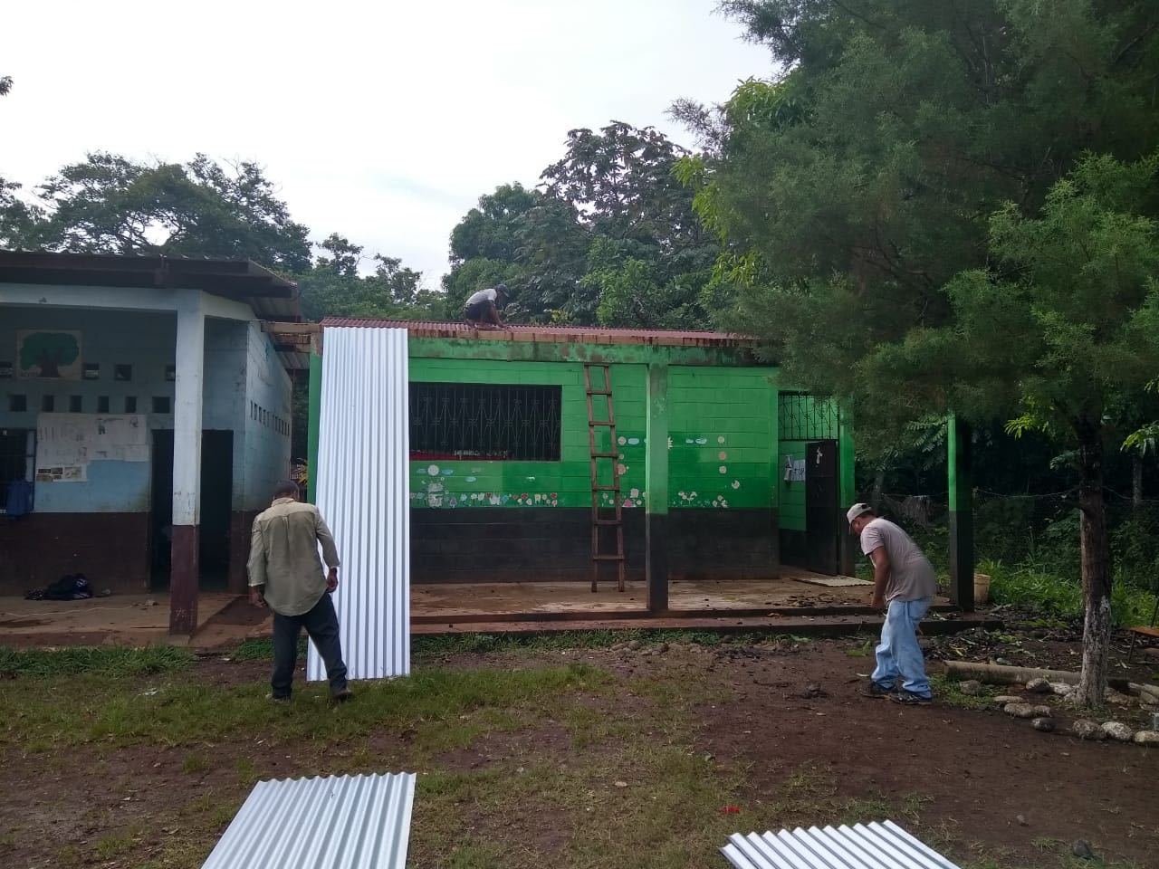 Three men replace the roof of a green classroom; one carries a new metal sheet while another works on top