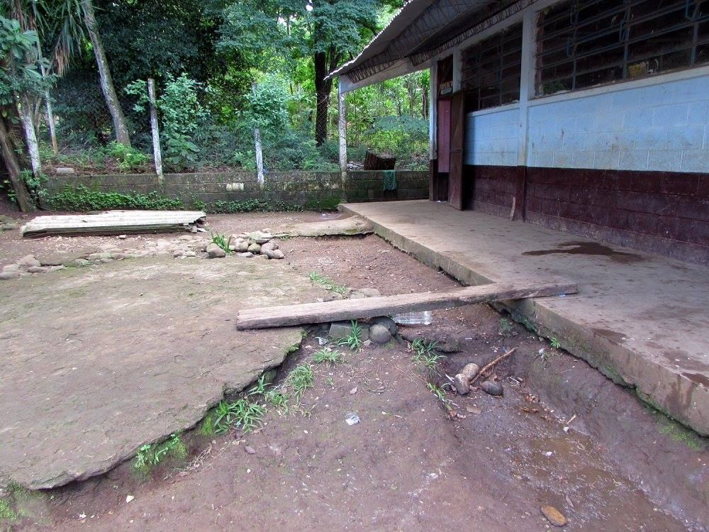 A wooden plank bridges a muddy ditch in front of a classroom corridor, next to a cracked concrete slab