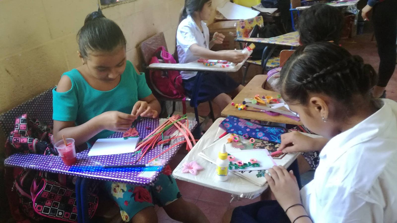 Girls in a classroom make cards decorated with rolled paper flowers, with scissors and glue on their desks