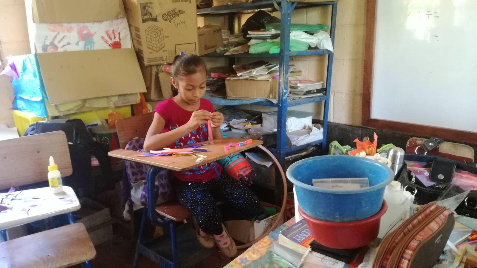 A girl rolls strips of paper at her desk in a classroom with shelves, cardboard boxes and a whiteboard