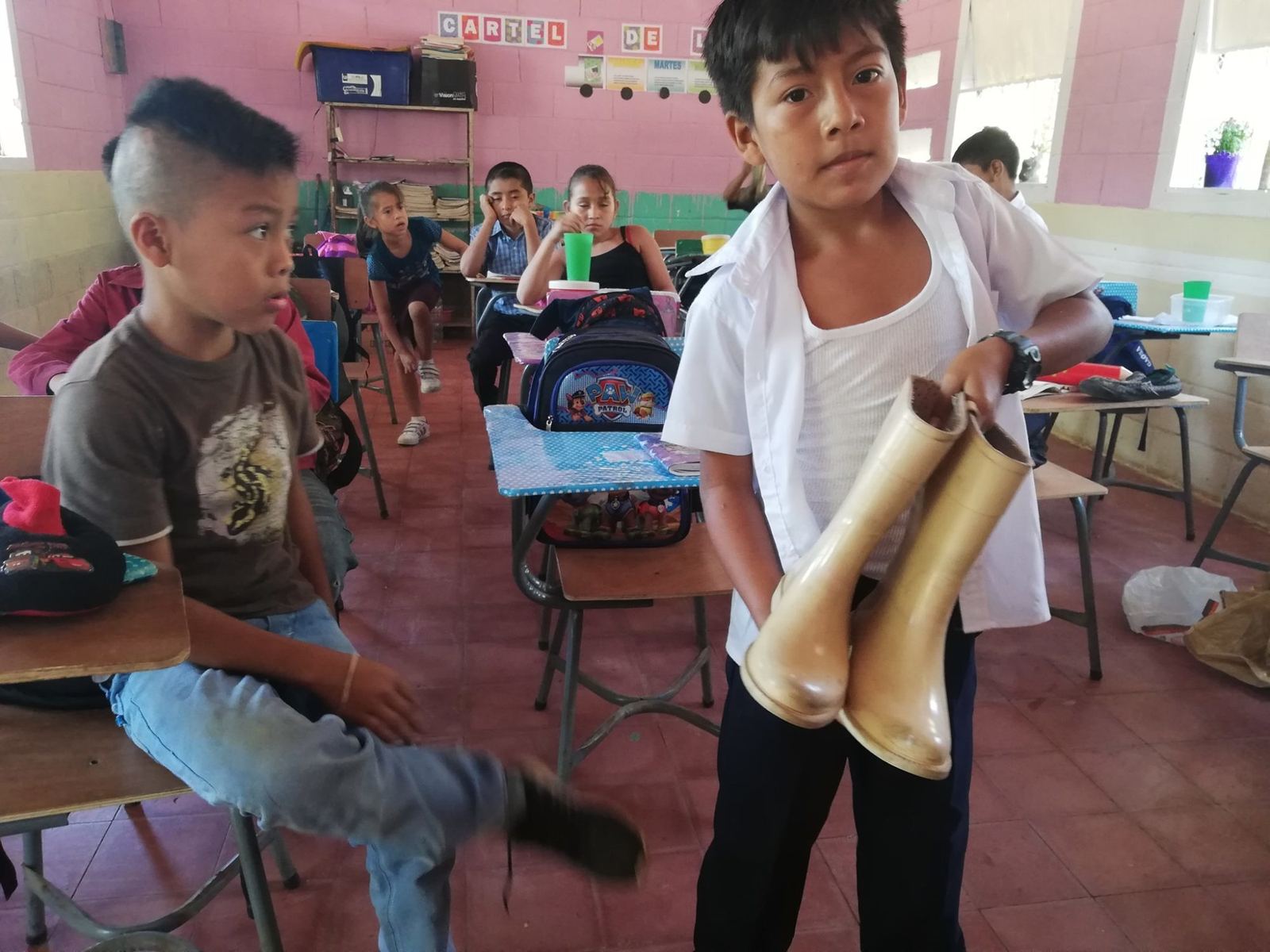 A schoolboy in uniform holds a pair of rubber boots in a classroom with pink walls