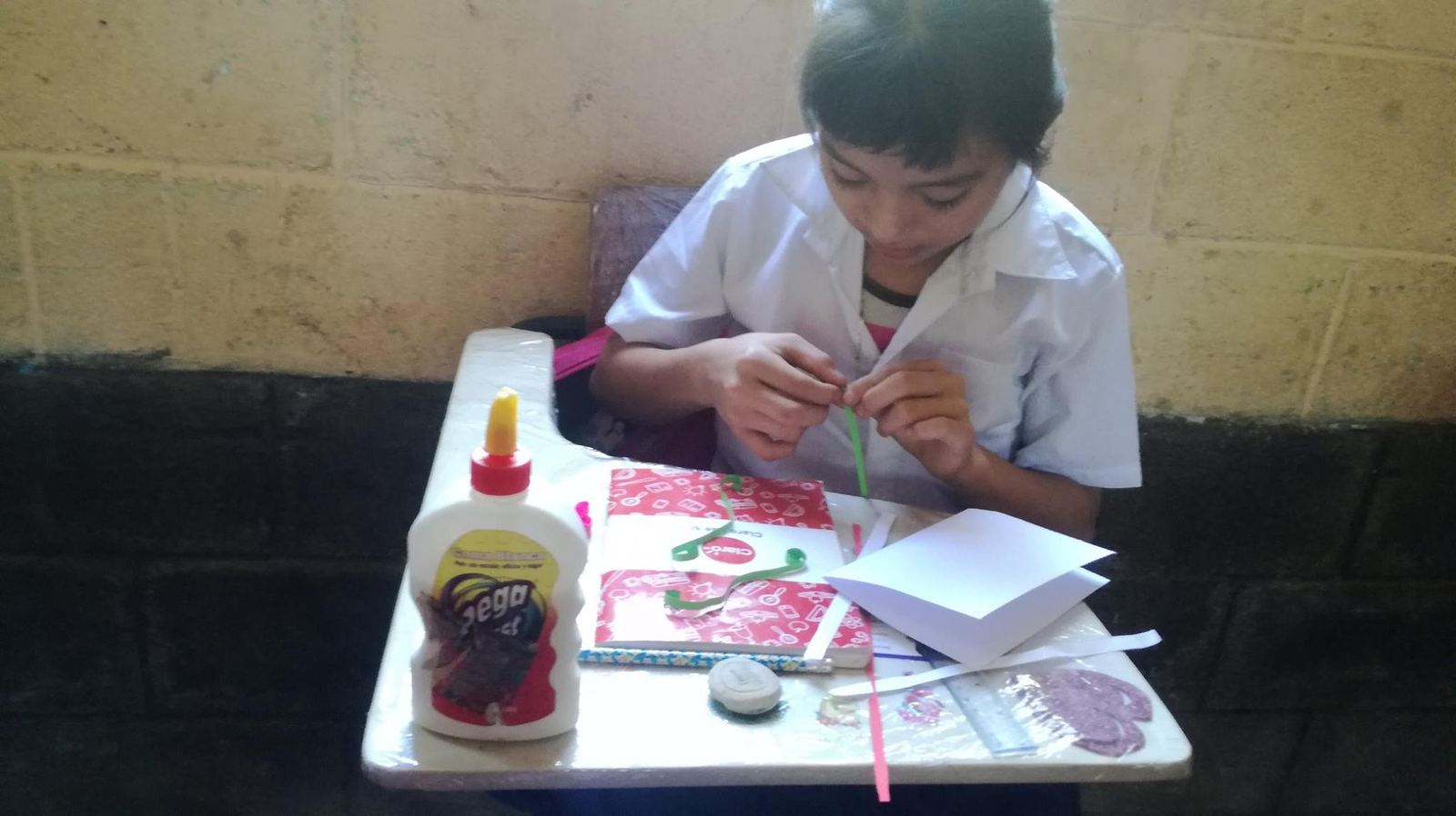 A student in uniform rolls a green paper strip at a desk beside a bottle of glue and a notebook