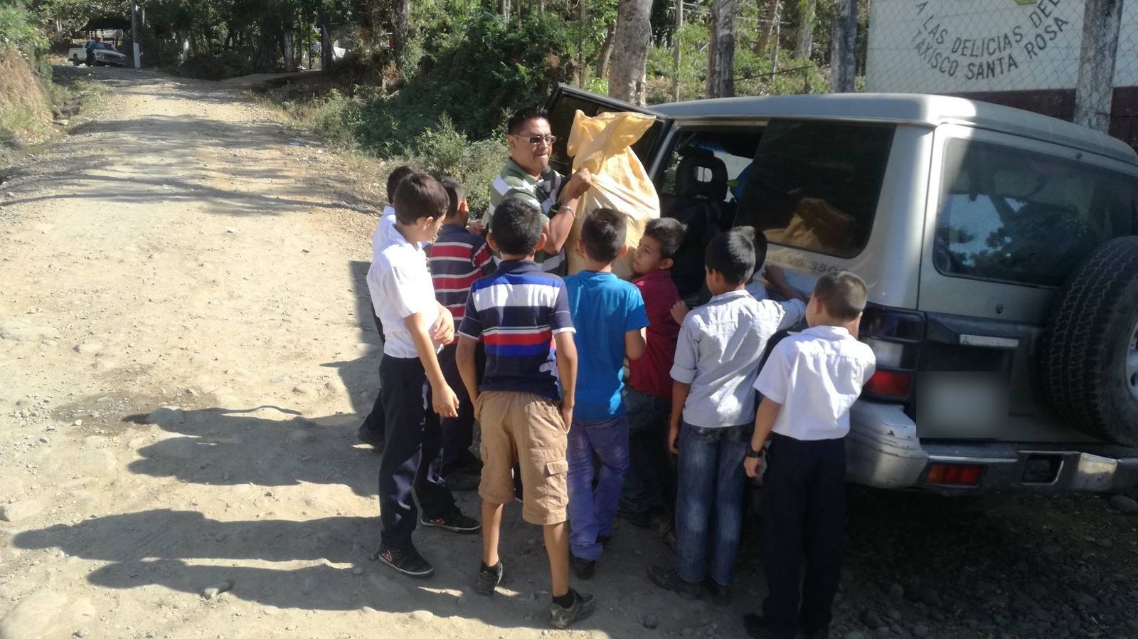 A man unloads a sack from an SUV, surrounded by boys in school uniforms on a dirt road