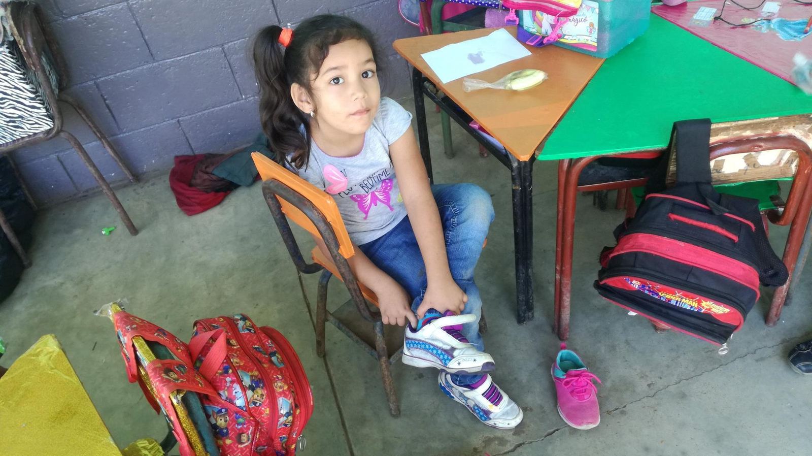 A little girl sits on a small chair putting on white sneakers, next to backpacks and a pair of pink sneakers