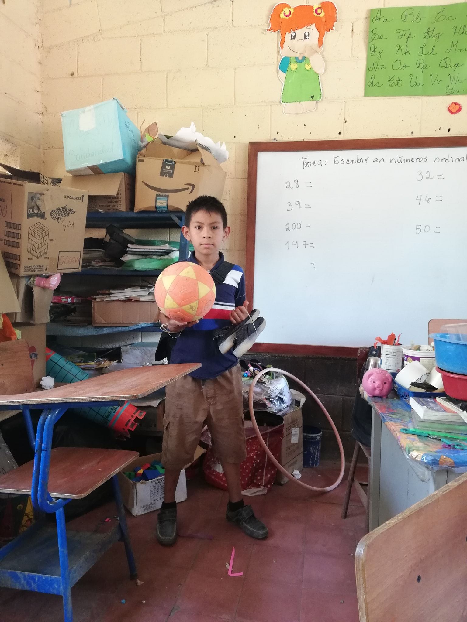 A boy holds a ball and a pair of sneakers in a classroom, in front of a whiteboard with a math assignment