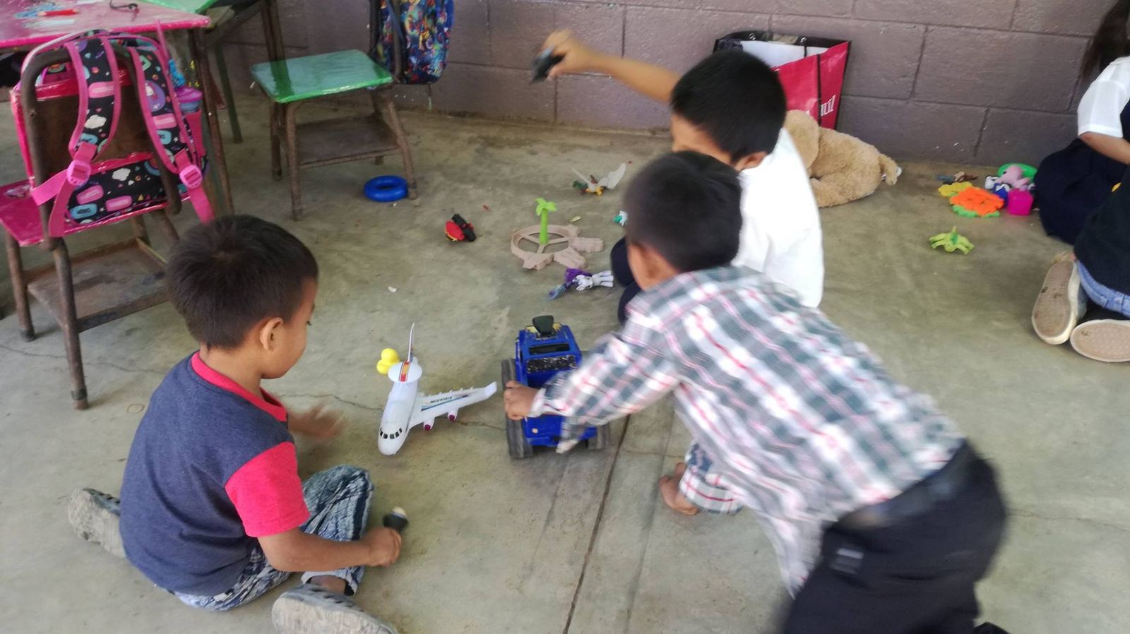 Three boys play on a concrete floor with a toy airplane, a blue toy vehicle and figurines