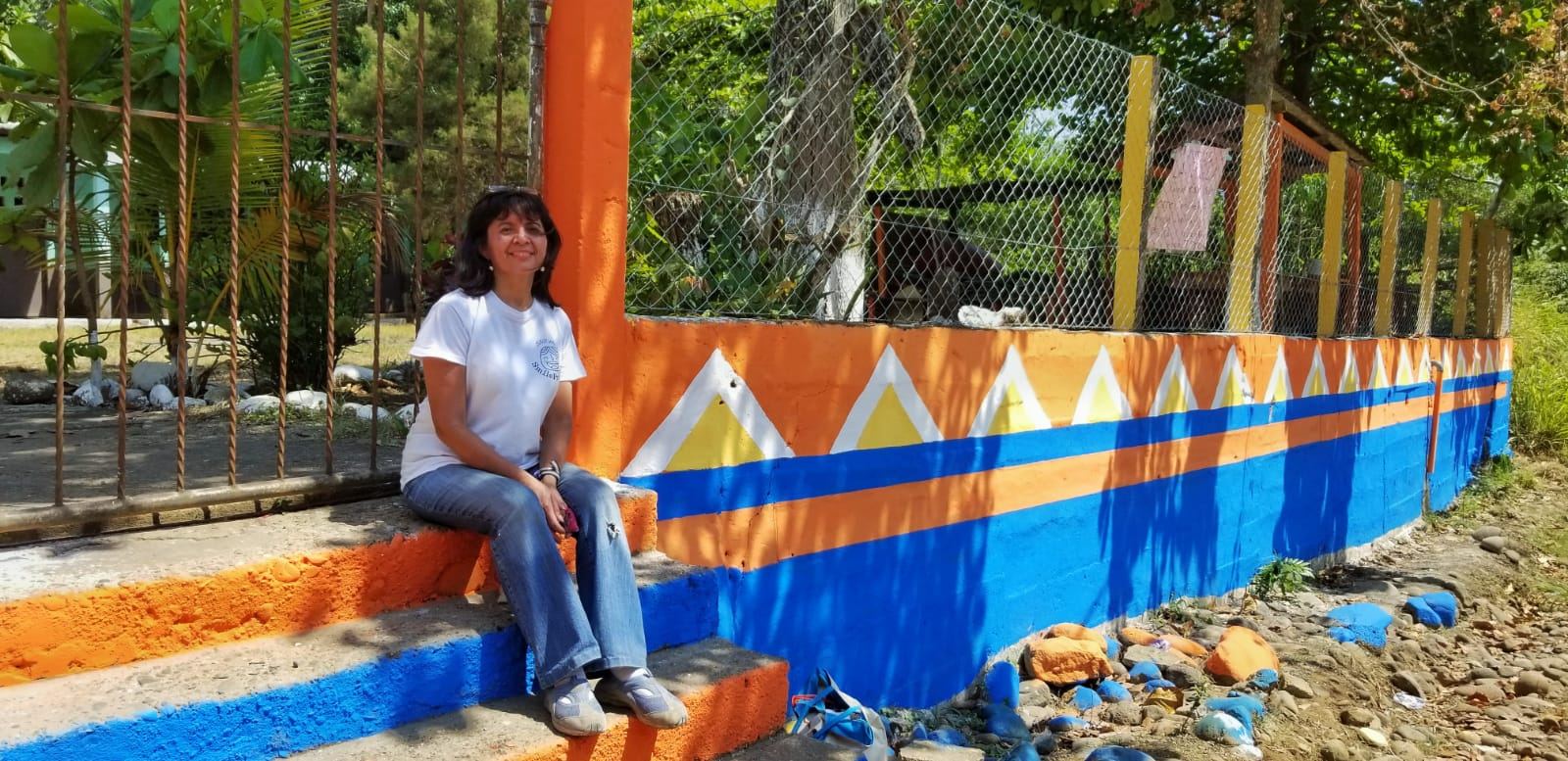 A smiling woman sits on the steps beside the school wall, freshly painted orange, blue, white and yellow
