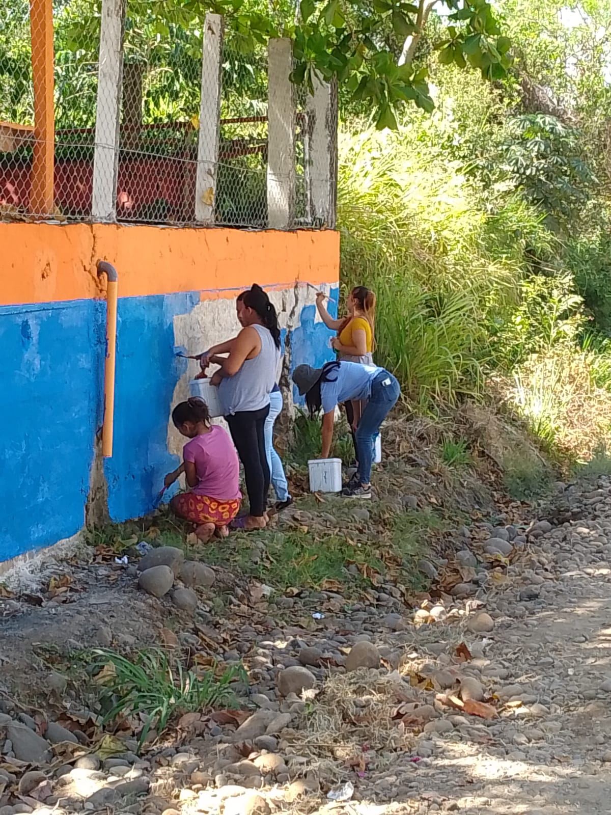 Four women and a girl paint the lower part of the school wall blue beside a dirt road