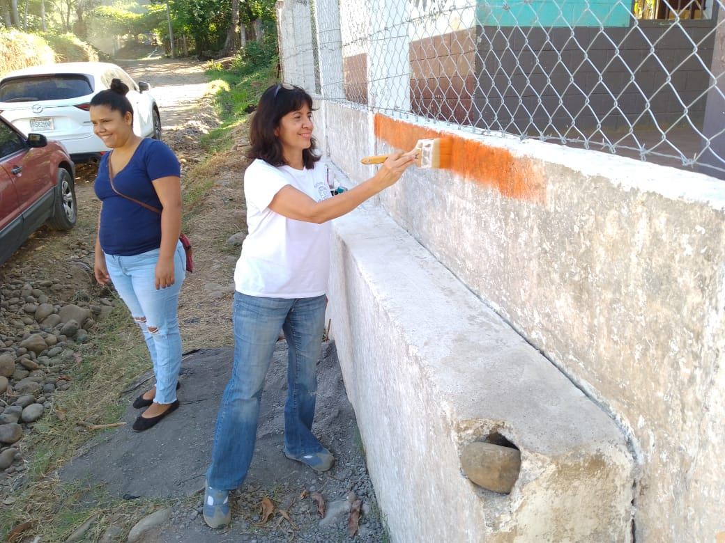 A woman brushes an orange stripe onto the school’s gray wall while another woman watches, smiling