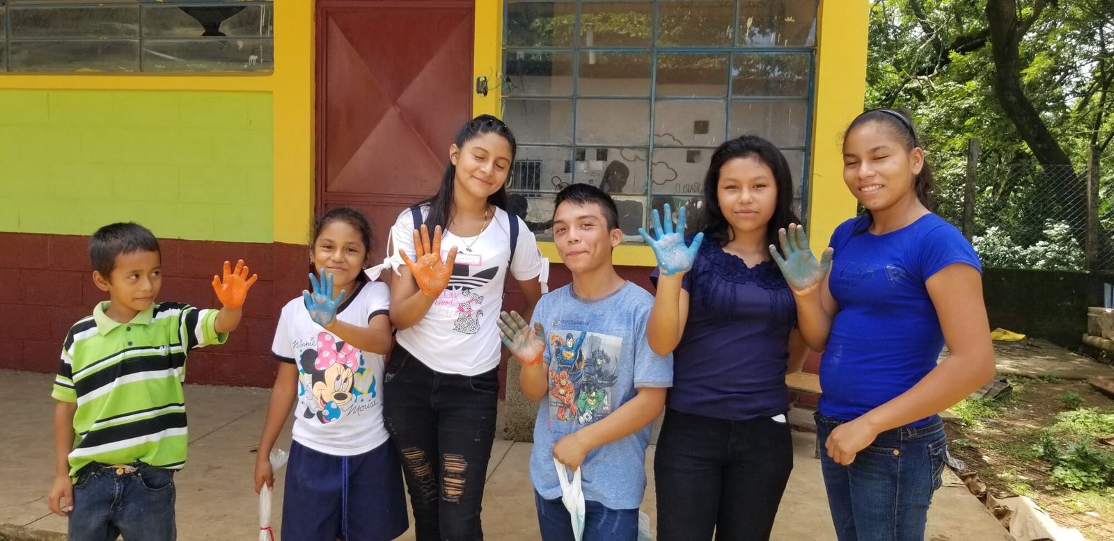 Six children and teenagers smile in front of a classroom, showing hands covered in orange and blue paint