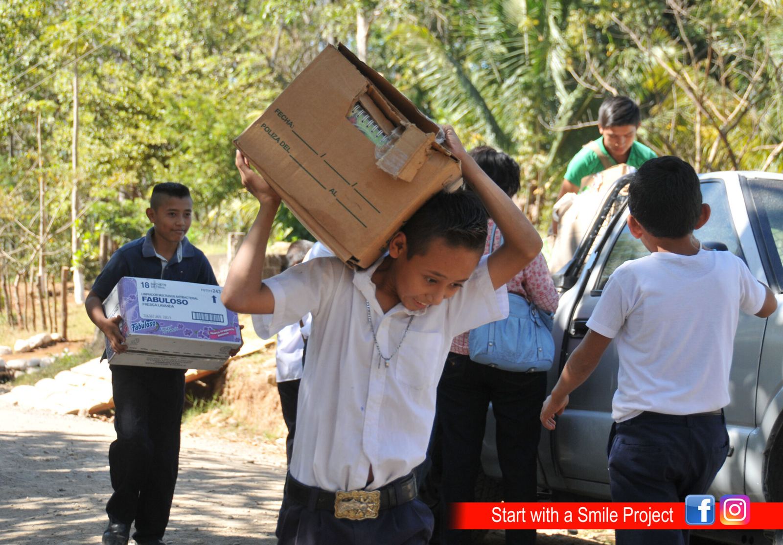 Uniformed schoolchildren carry boxes of notebooks and cleaning supplies from a pickup truck