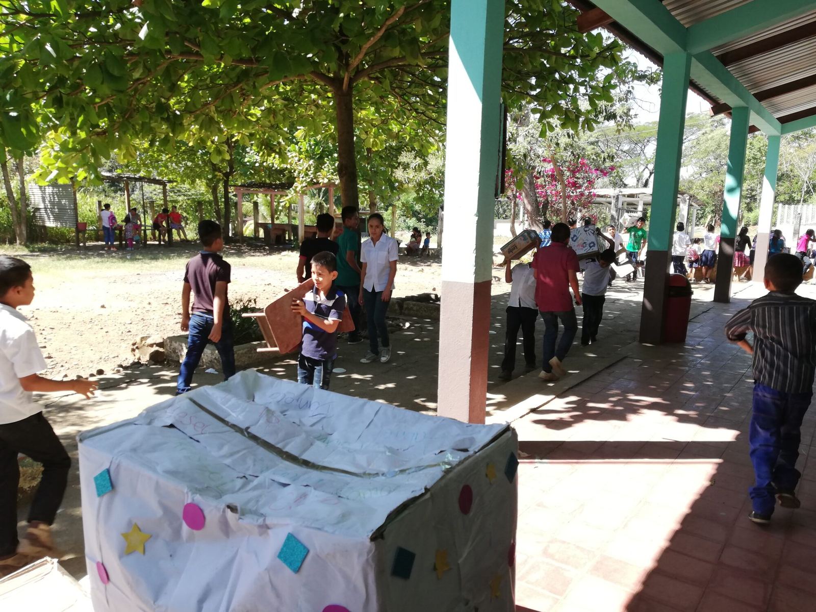Uniformed children carry boxes and a small table across the school courtyard beside a turquoise-pillared corridor
