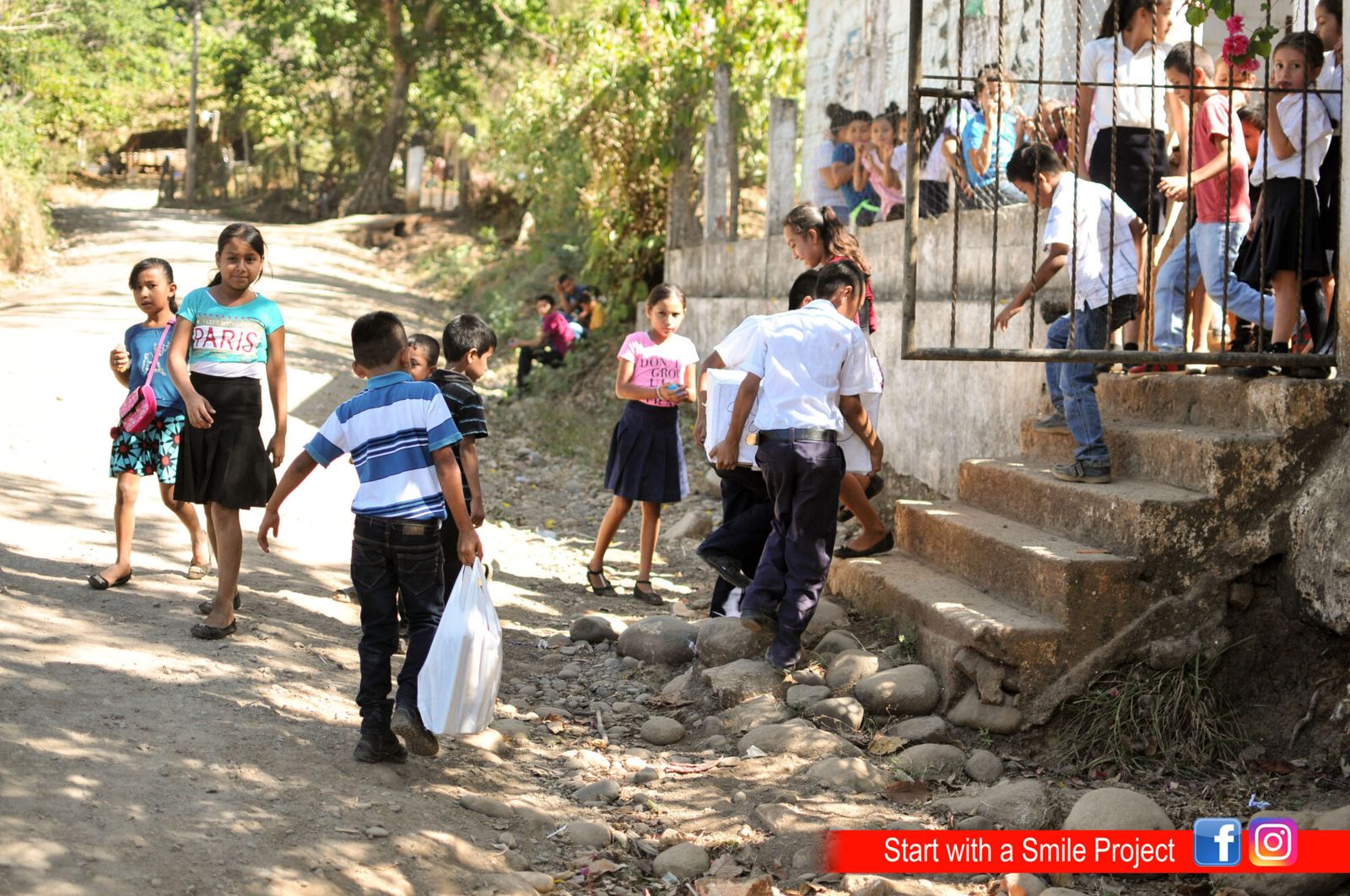 Two students carry a box up the school steps as other children watch from the gate