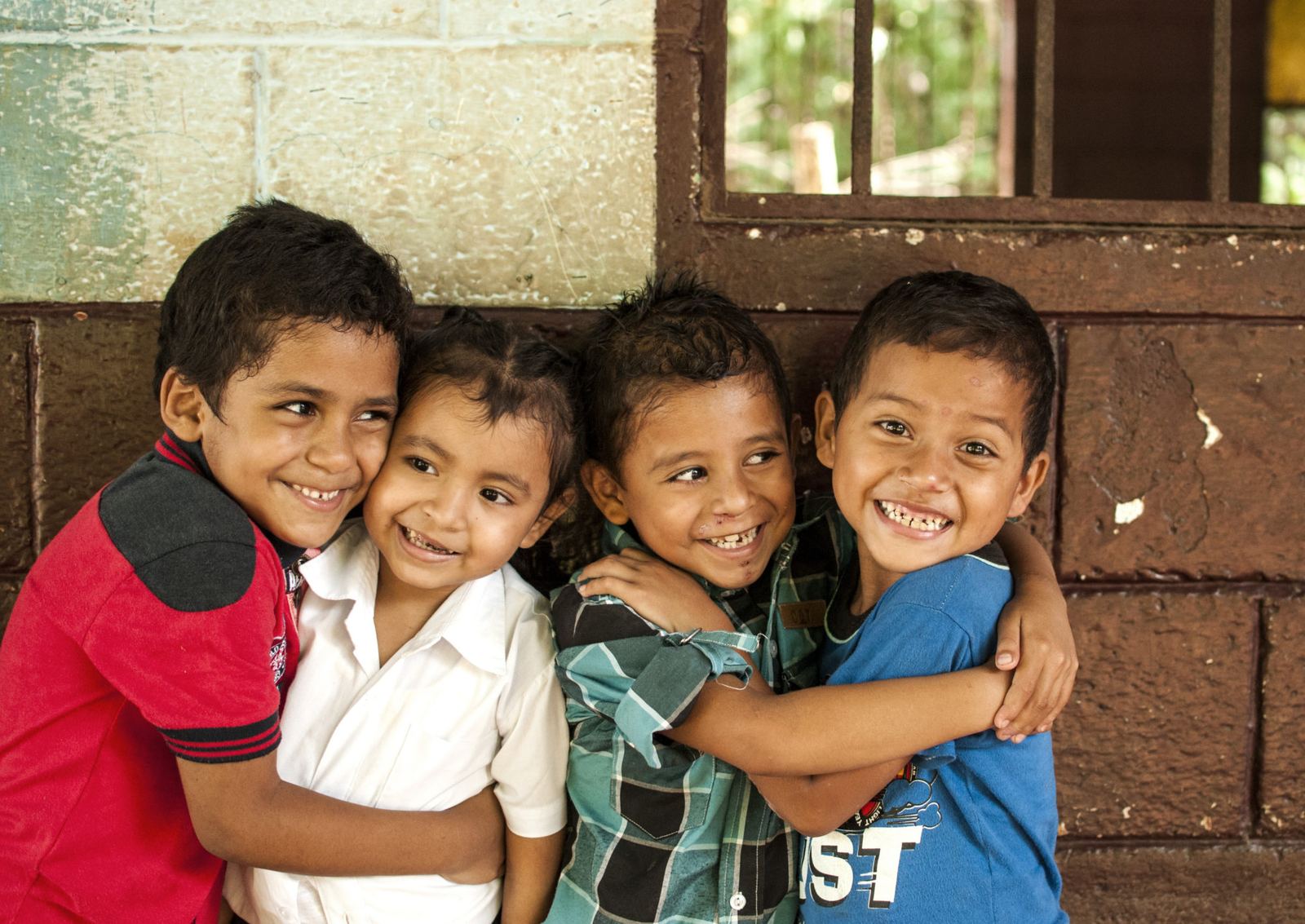 Four young children hug and laugh in front of a block wall beside a barred window