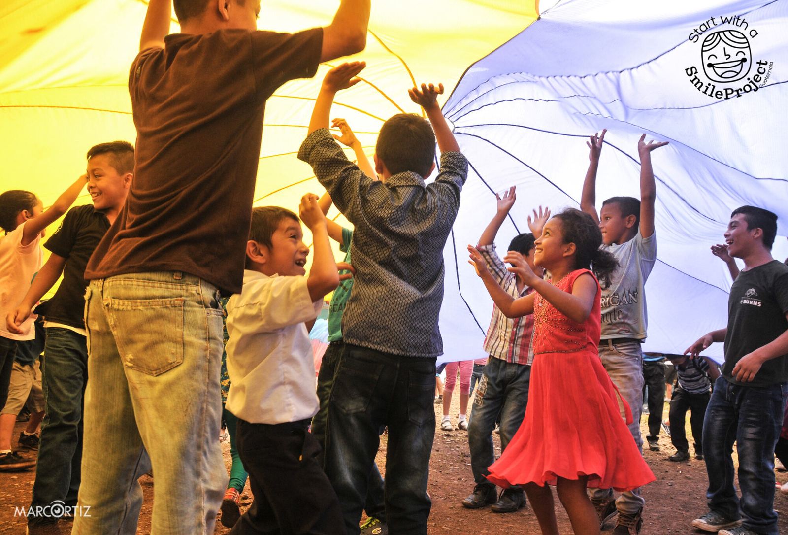 Girls and boys jump and laugh under a yellow and white play parachute