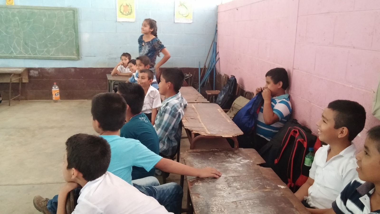 Students seated at worn wooden desks smile as they look toward the front of the classroom
