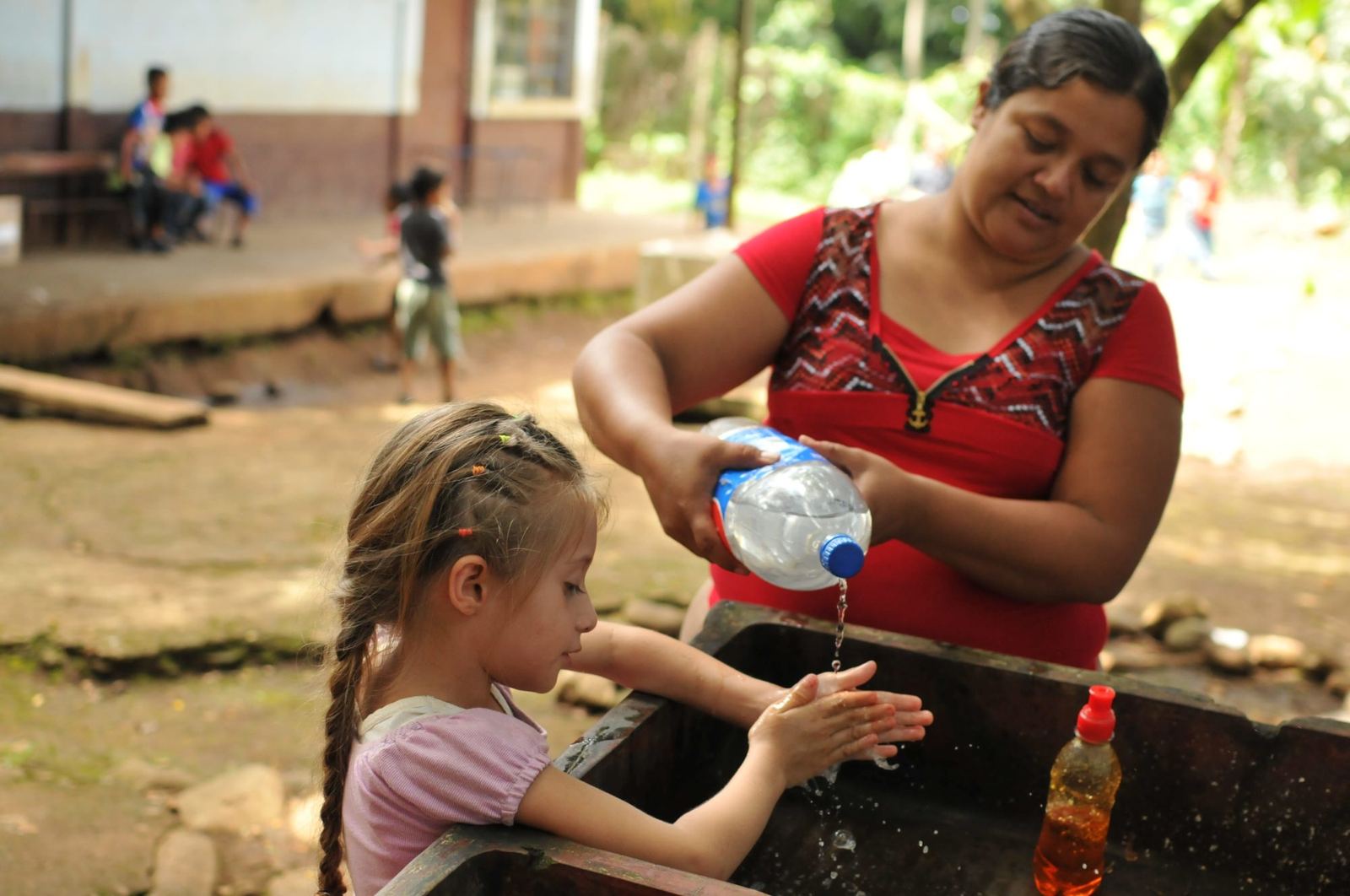 A woman pours water from a bottle so a young girl can wash her hands at an outdoor sink, soap beside her