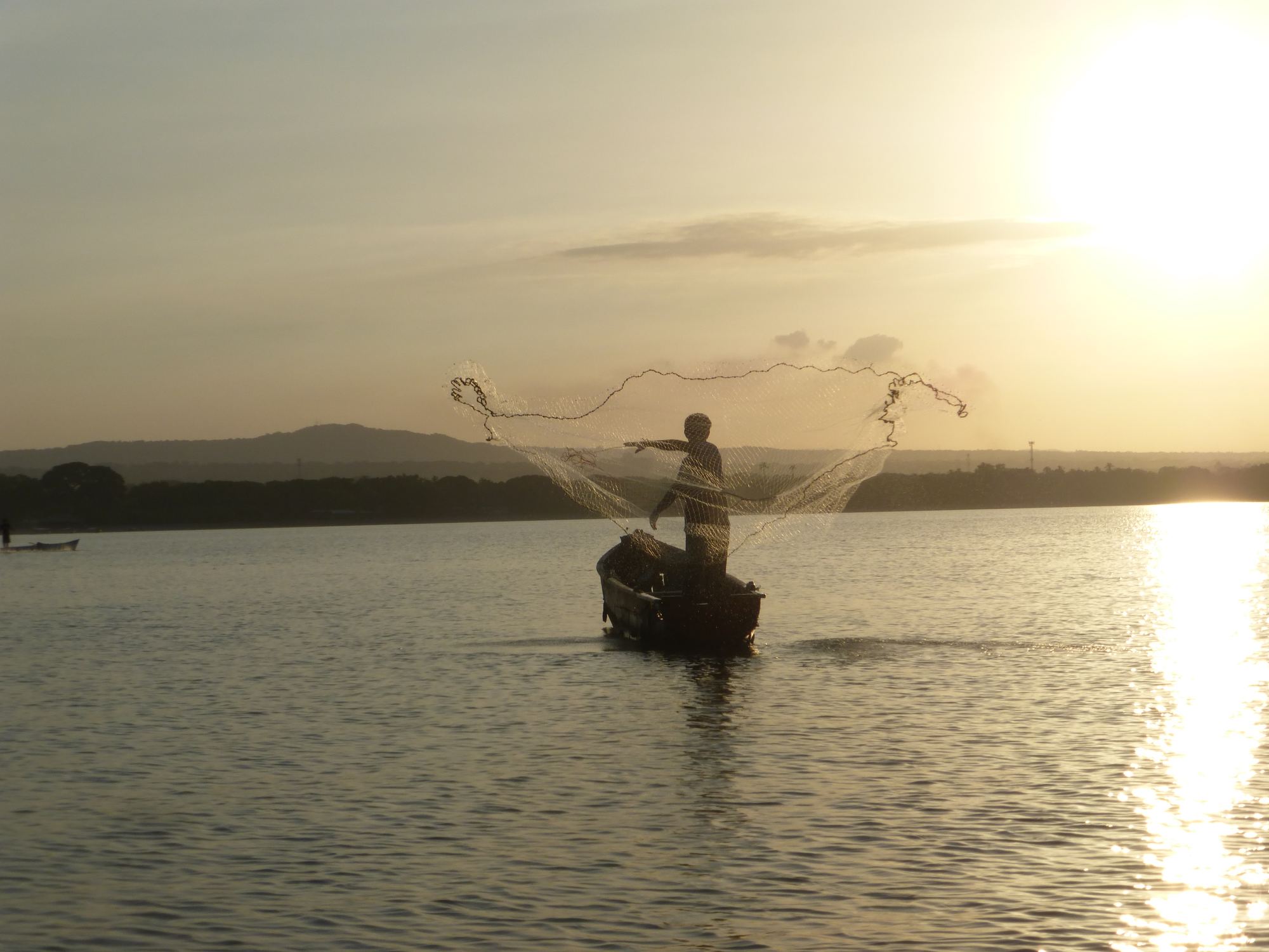 A fisherman casts his net from a small boat, backlit by the low sun over calm water