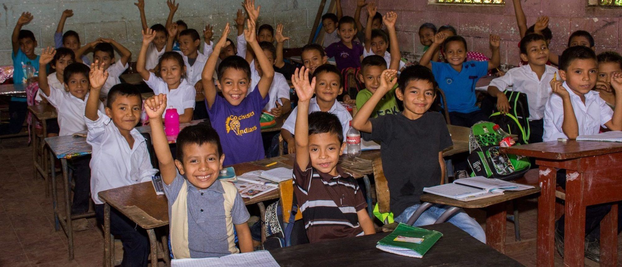 Niñas y niños de primaria sonríen con los brazos en alto en un aula llena de pupitres de madera