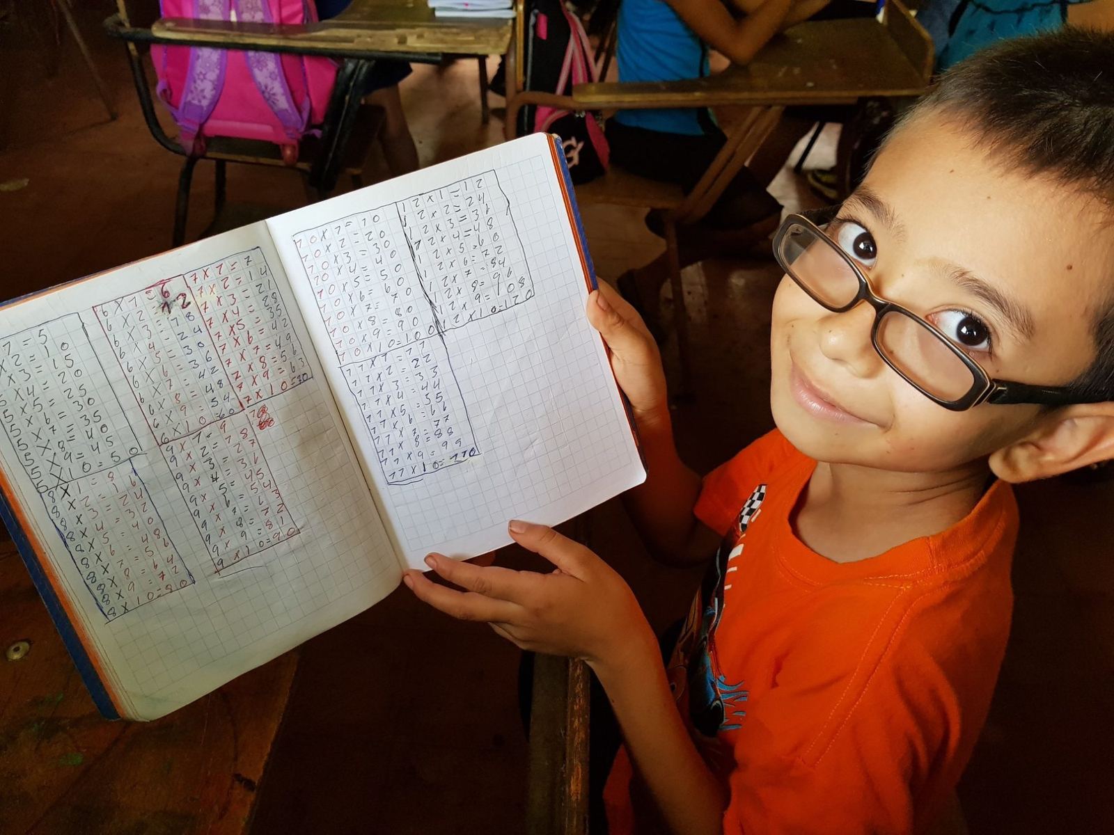 A boy in glasses proudly shows his notebook of multiplication tables in the classroom