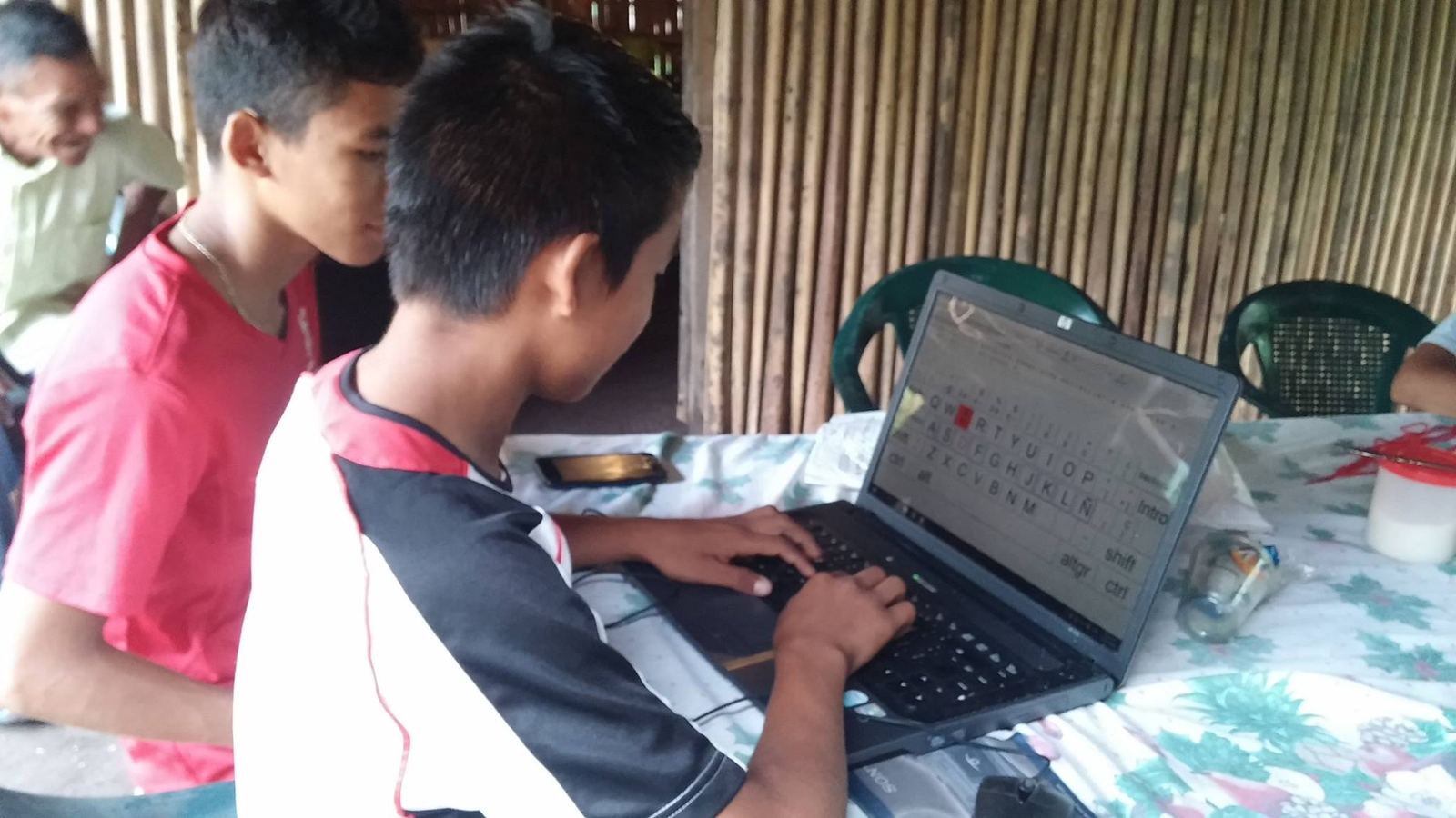 Two teenage boys practice typing on a laptop in a room with bamboo walls