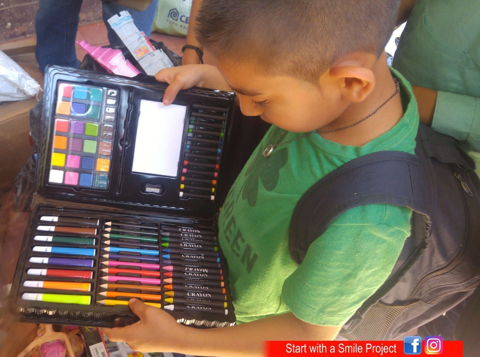 A boy with a backpack looks through an art kit with watercolors, markers, colored pencils and crayons