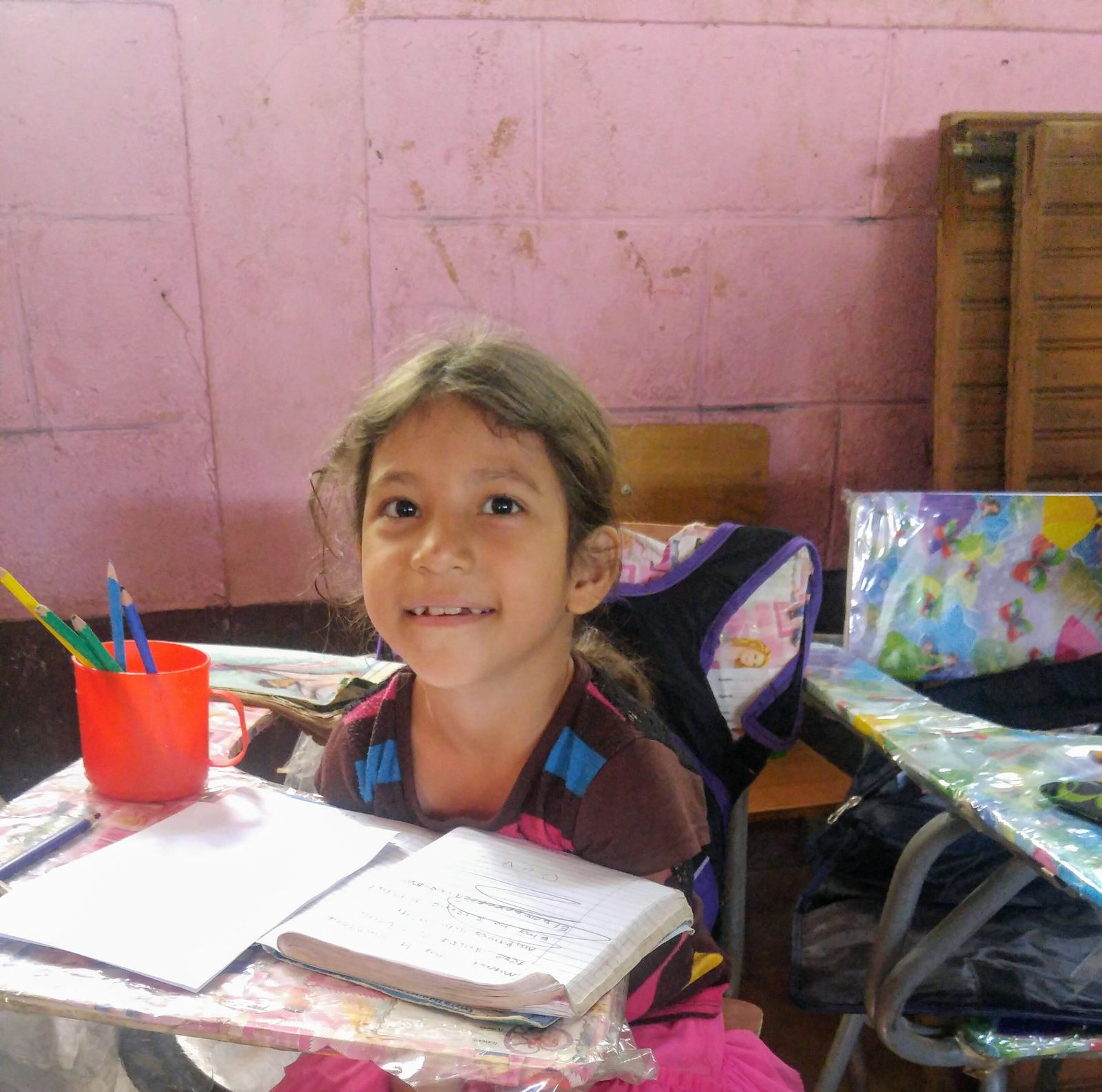 A girl smiles at her desk beside her notebook and a red cup of colored pencils, in front of a pink wall