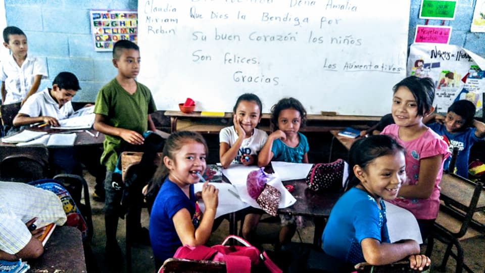 Children smile in their classroom in front of a whiteboard with a thank-you message for Amalia