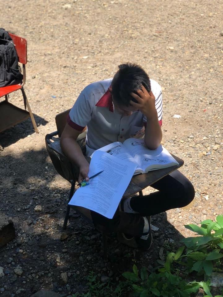 A uniformed student reads a textbook and a worksheet at an outdoor desk, seen from above