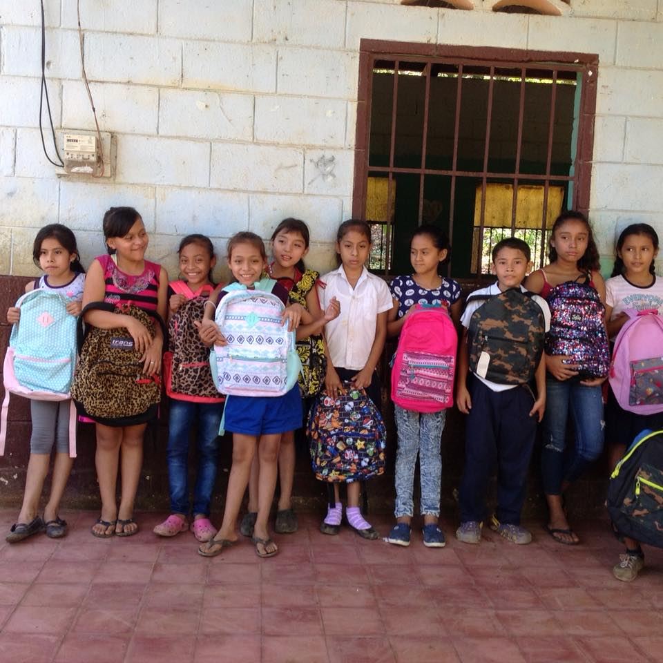 Ten children line up against a school wall, each holding a backpack