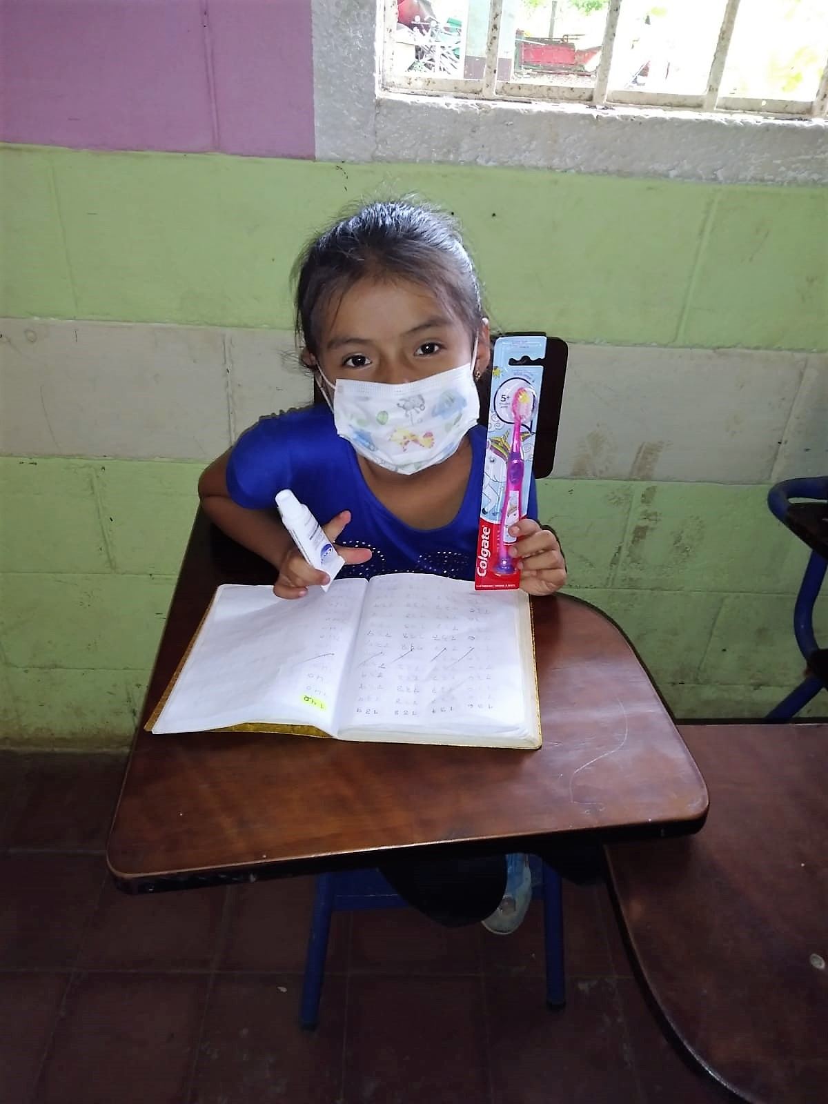 A girl in a face mask holds up a toothbrush and toothpaste at her desk, next to her open notebook