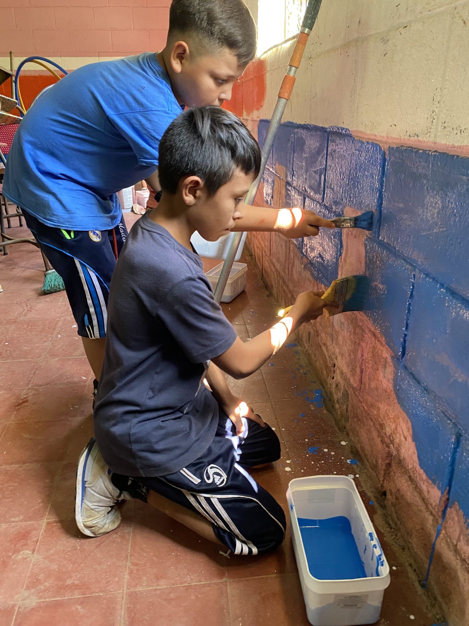 Two boys paint the lower part of a school wall blue with brushes and a tub of paint