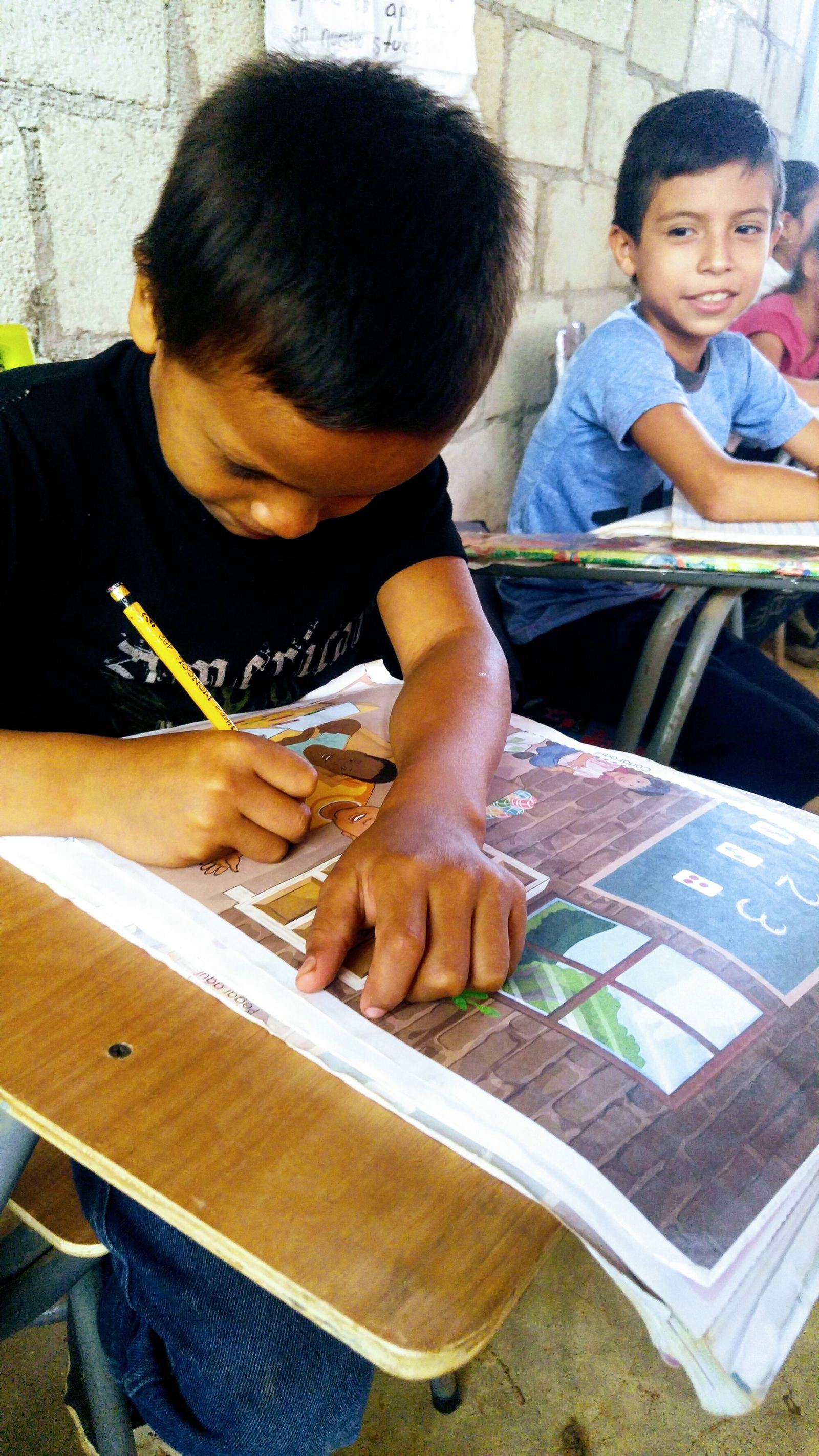 A boy colors in his workbook with a pencil while a classmate smiles at the camera