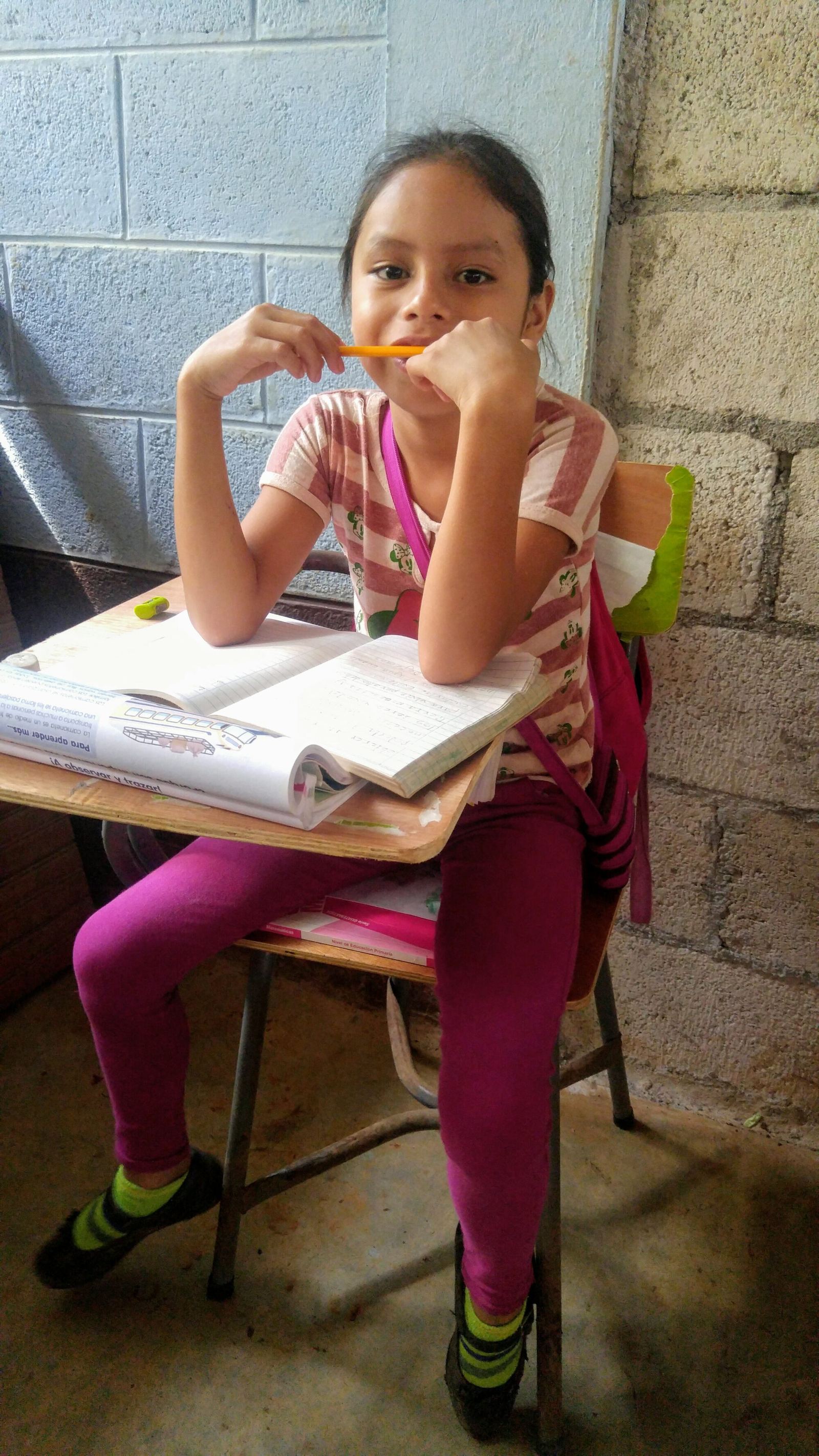 A girl poses at her desk with a pencil held to her lips, elbows on her notebook and textbook