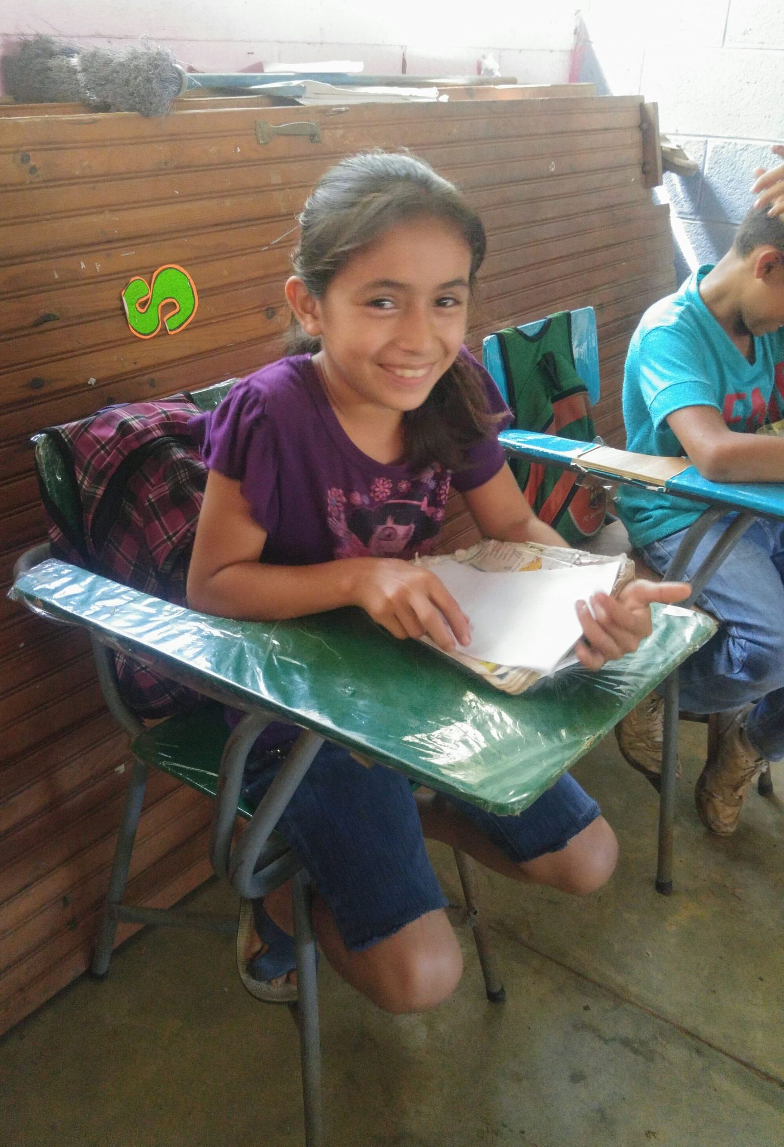 A girl smiles at her green desk, holding some sheets of paper, in front of a wooden panel